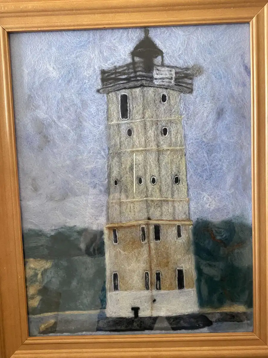 Needle felt portrait of a lighthouse with a gray-blue sky background and shrubbery covering the rocks.